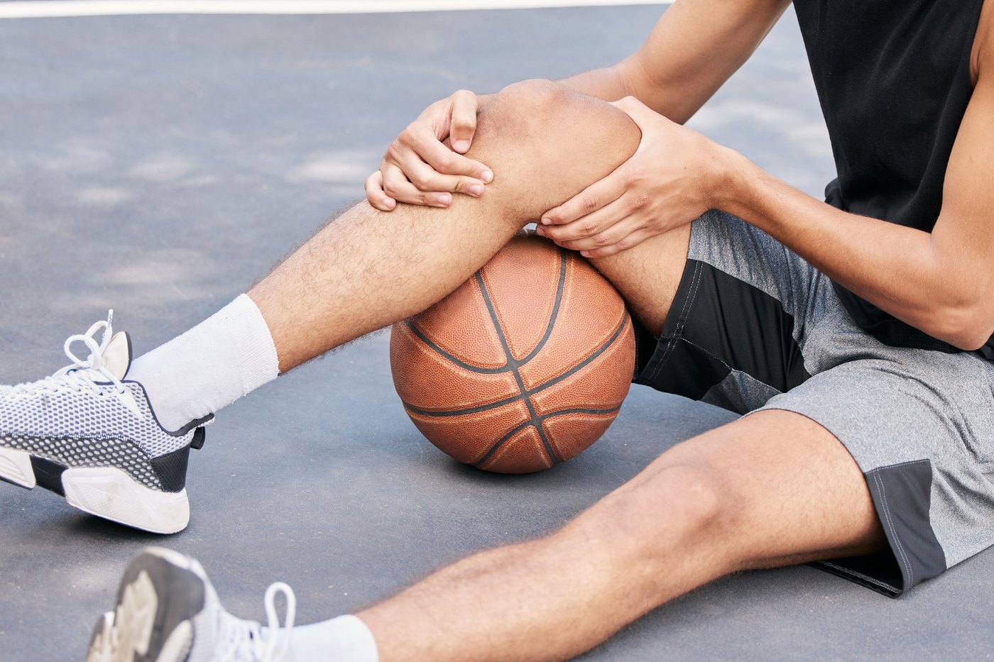A basketball player sitting on a court holding his knee while another person supports the leg