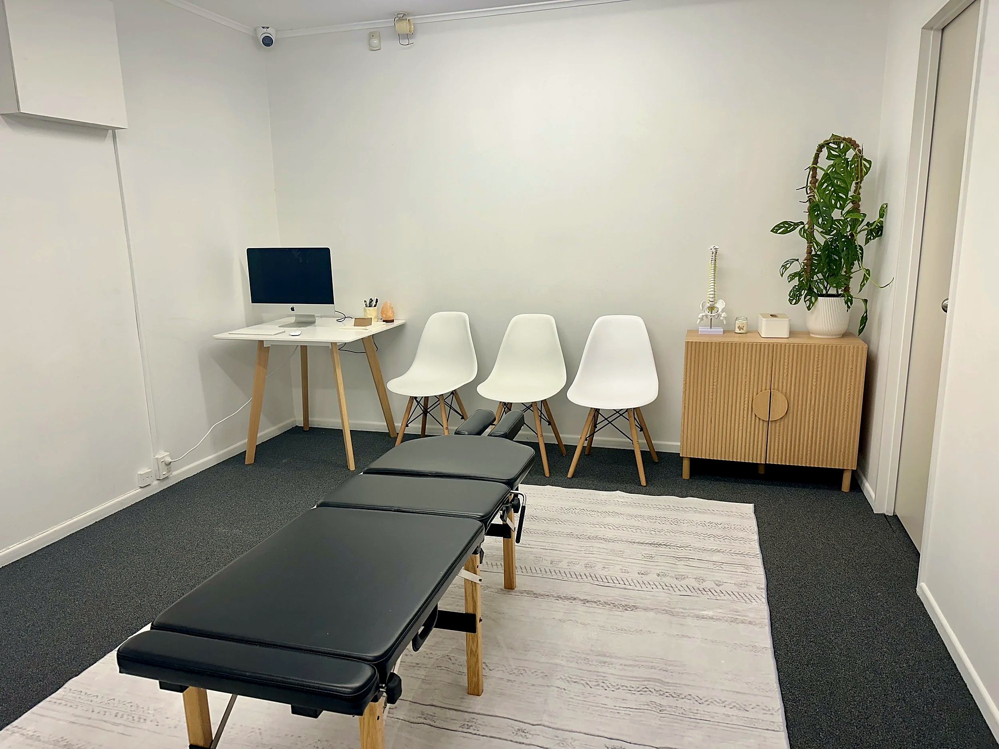 A treatment room at The Healing Centre Henderson: adjusting table, white chairs, desk and a plant